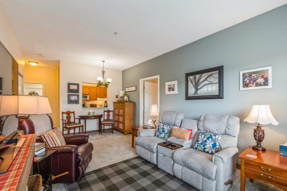 A cozy and well-decorated living room featuring comfortable seating, a rug, and framed artwork on the walls, with an adjacent dining area in the background.