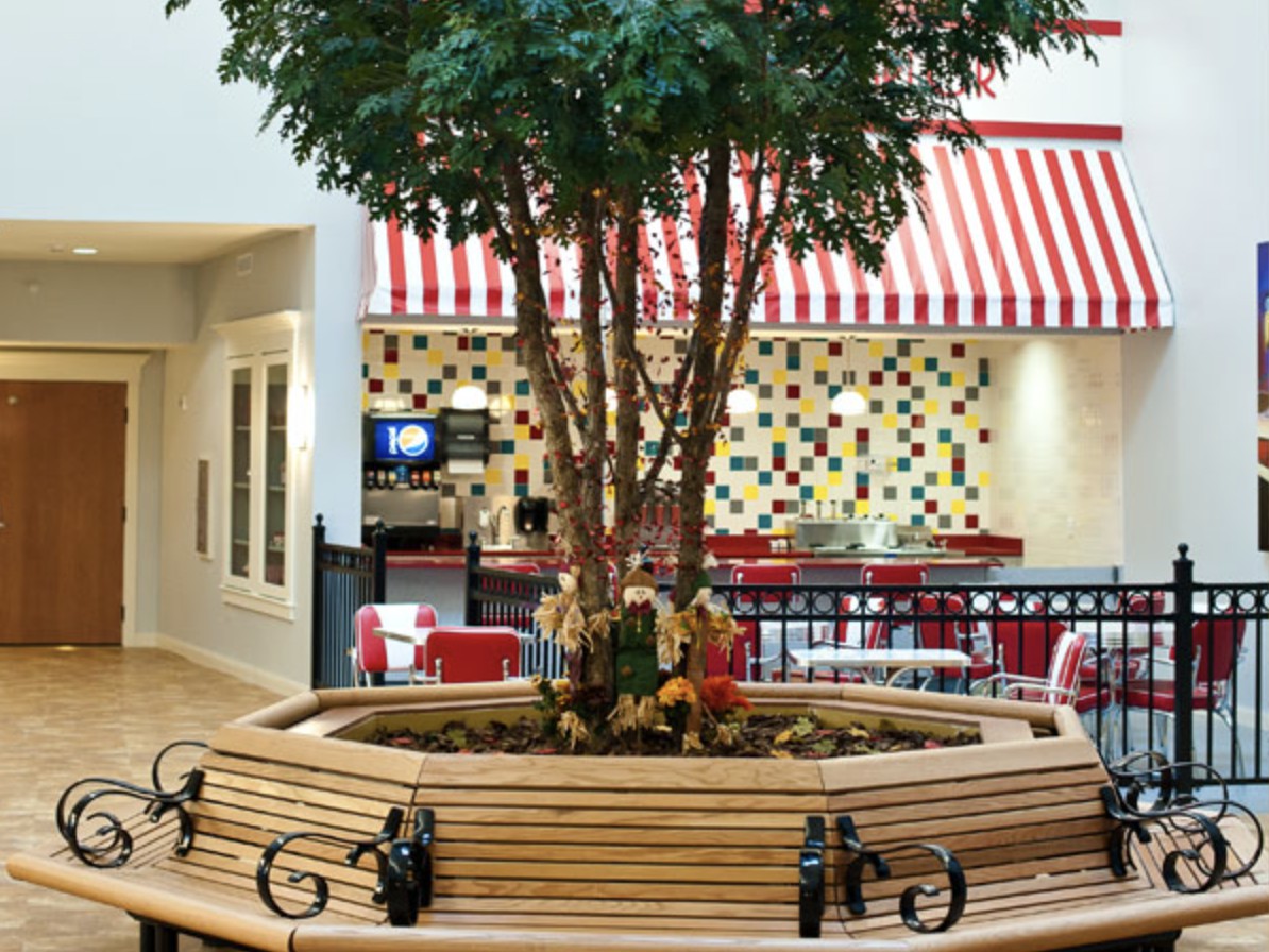 A cozy indoor seating area with benches surrounding a central tree in a mall's food court, featuring striped awnings and colorful tiles.