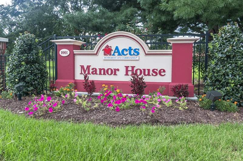 Entrance sign for the Acts Manor House surrounded by landscaped flowers.