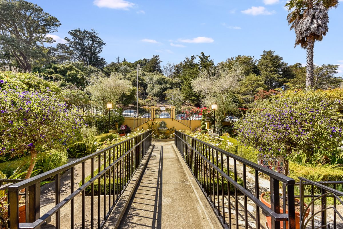 A scenic pathway bridge leading through a lush garden filled with trees and greenery under a clear blue sky.