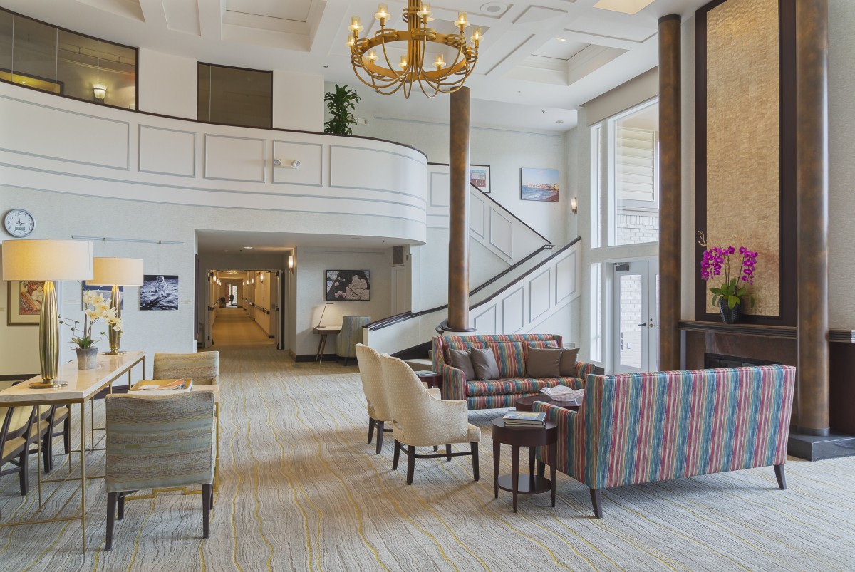 A spacious hotel lobby with a seating area, elegant chandelier, and stairway leading to an upper level.