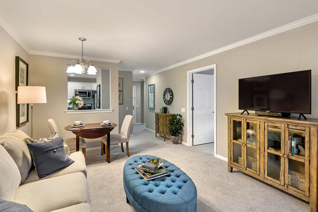 A cozy living room with a dining area, featuring a sofa, a tufted ottoman, and a wooden TV stand.