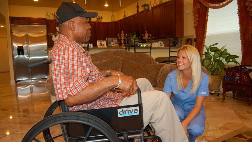 A caregiver kneels next to a man in a wheelchair, engaging in conversation.