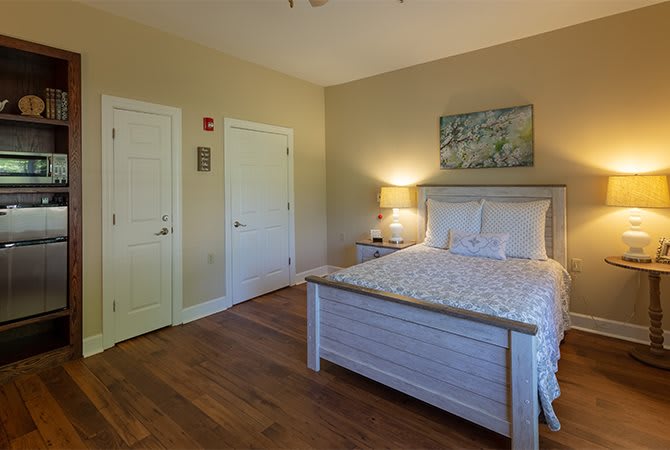 A cozy bedroom with a wooden floor, featuring a neatly made bed, a bedside lamp, and an artwork hanging on the wall.