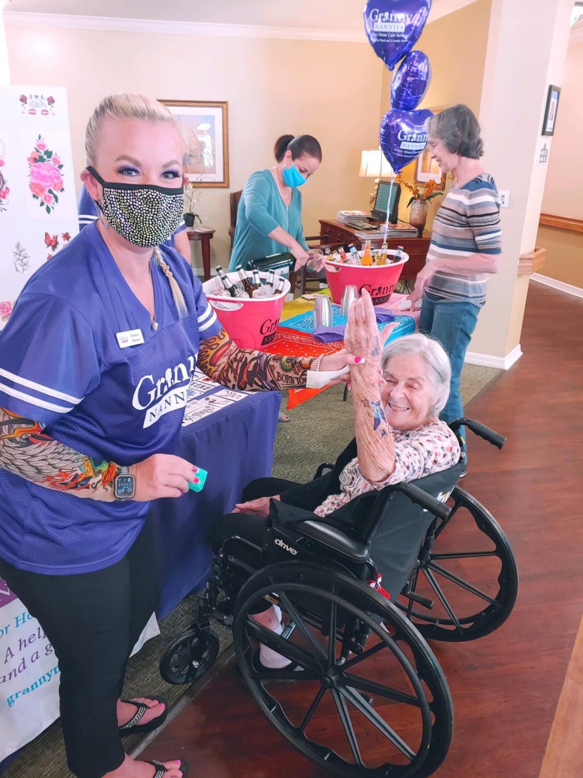A healthcare worker and an elderly woman in a wheelchair celebrate together, surrounded by party decorations indoors.