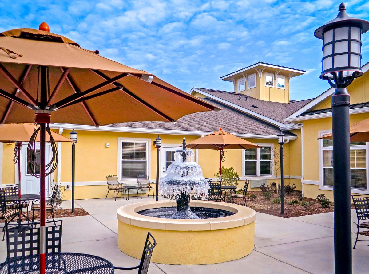 A sunny courtyard with a central fountain, surrounded by yellow buildings and outdoor seating with umbrellas.
