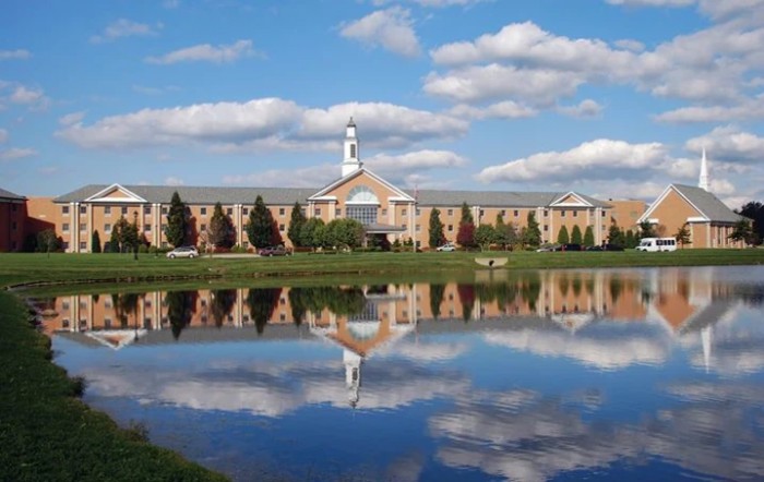 A large building reflected in a calm lake under a partly cloudy sky.
