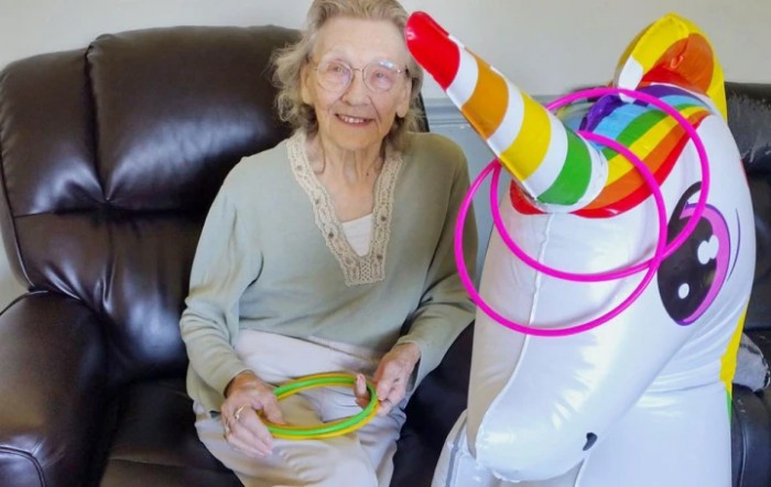 An elderly woman is sitting on a couch, smiling, with a colorful inflatable unicorn and hula hoops.