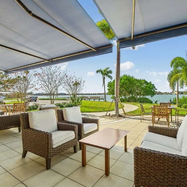 A serene patio with comfortable wicker seating overlooks a tranquil lake surrounded by lush greenery and palm trees.