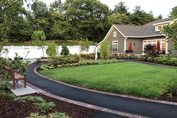 A well-maintained garden with a lush lawn, trees, and a neat driveway in front of a house.