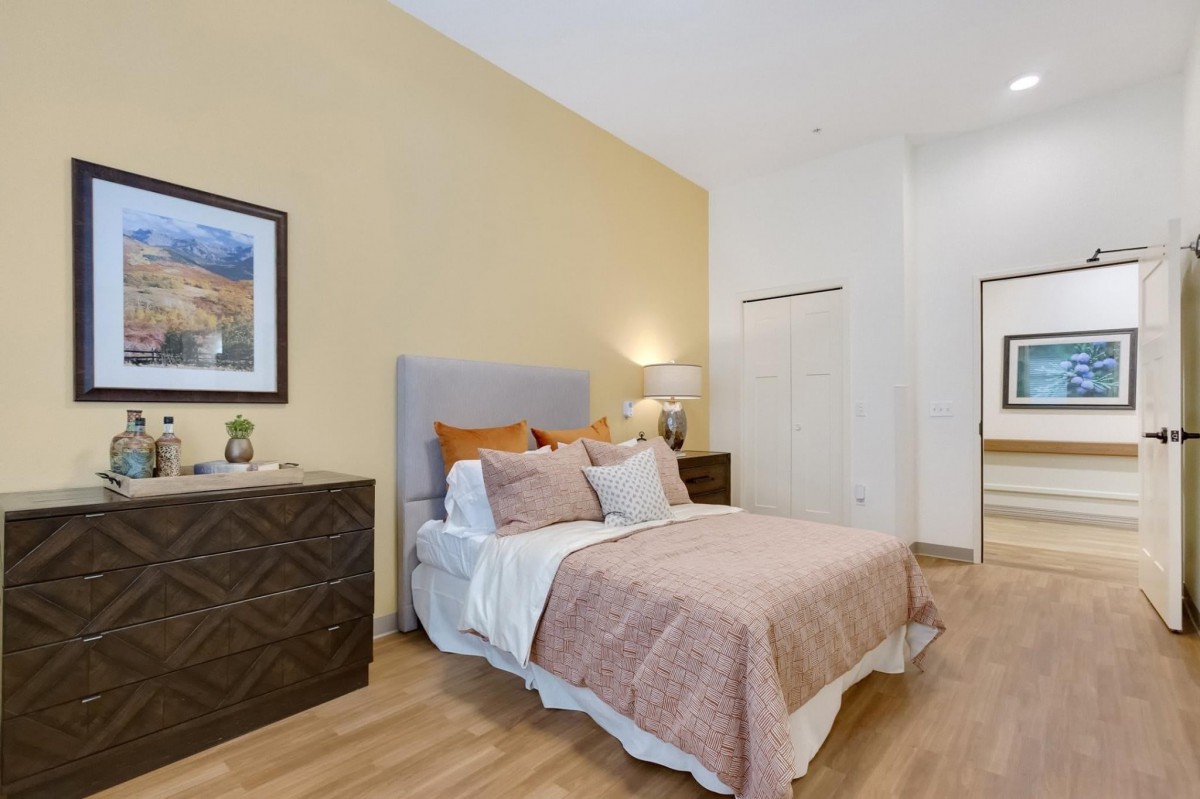 A cozy bedroom featuring a neatly arranged bed with modern bedding, framed artwork on the wall, and stylish furnishings including dressers and a lamp.