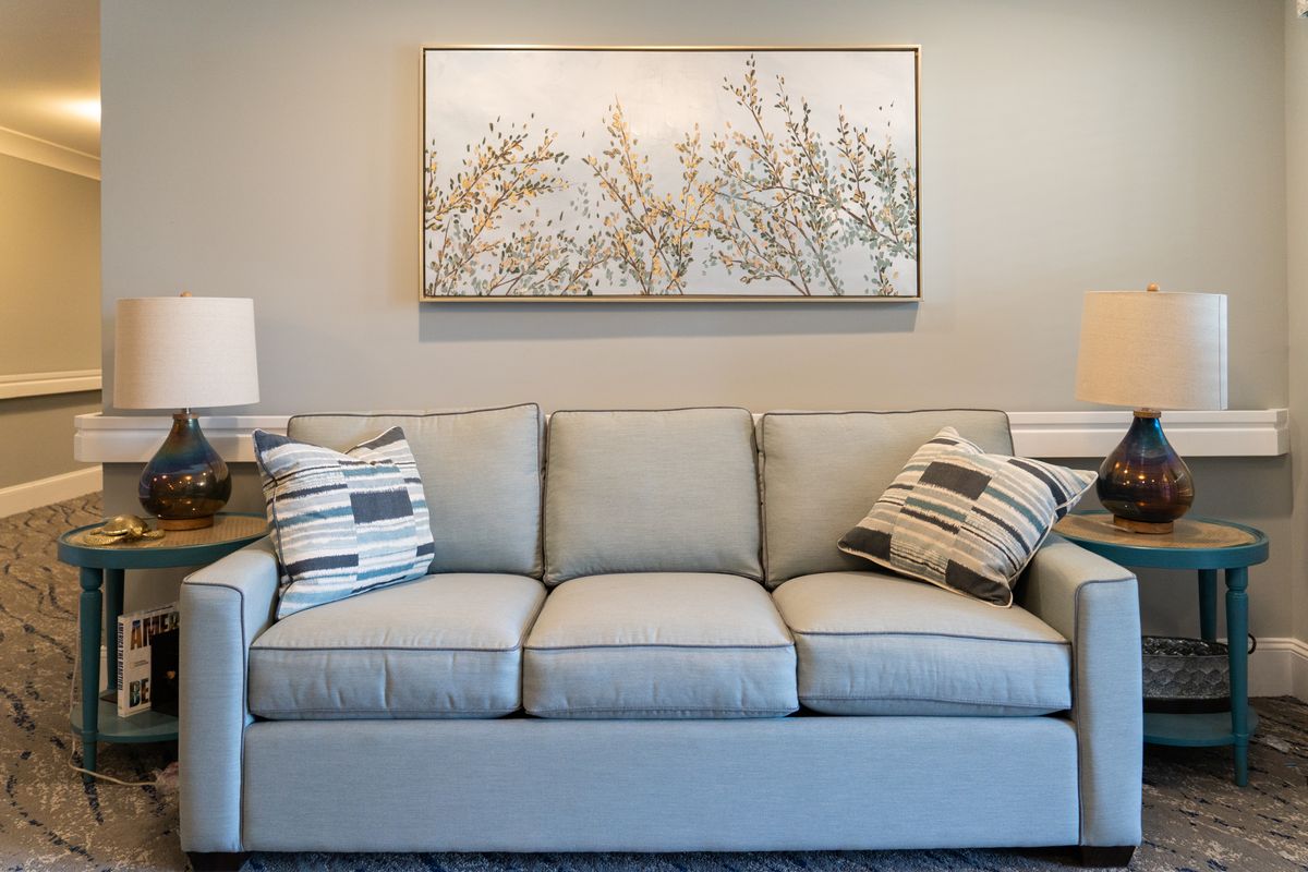 A cozy living room featuring a light gray sofa with patterned cushions and a nature-inspired artwork above.
