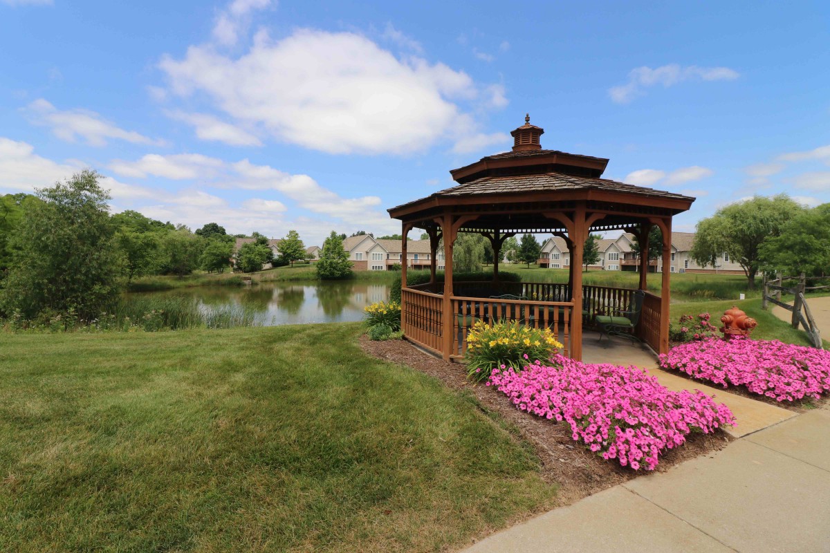 A scenic view of a wooden gazebo by a lake with colorful flowers in a park.
