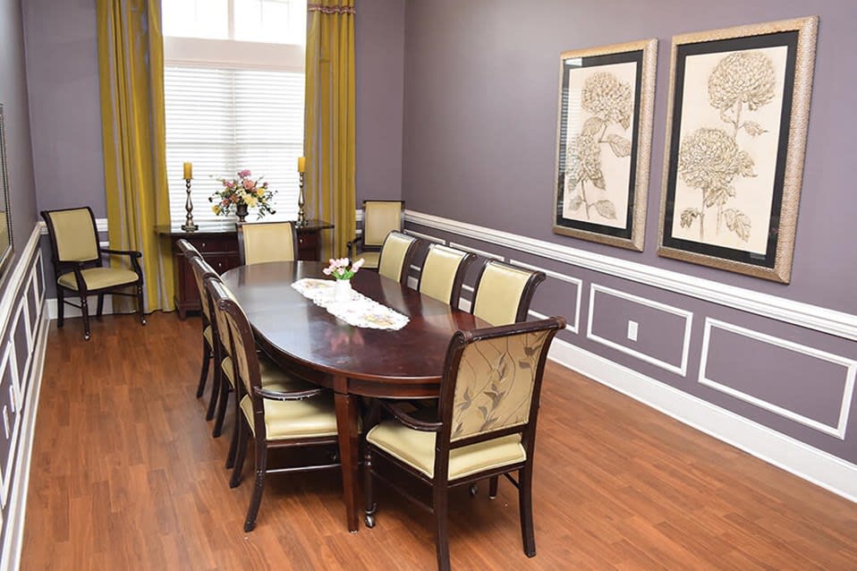 A well-decorated dining room featuring a long wooden table with upholstered chairs, framed artwork on lavender walls, and hardwood flooring.
