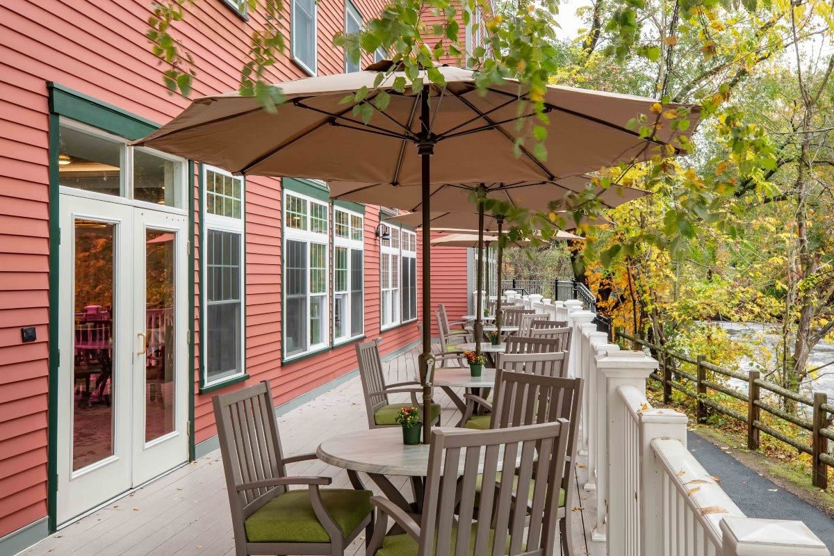 A cozy outdoor patio with tables and umbrellas on a terrace beside a red building, surrounded by lush greenery.