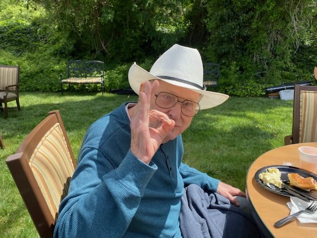 An elderly man wearing a cowboy hat and glasses makes an 'OK' gesture while sitting outdoors.