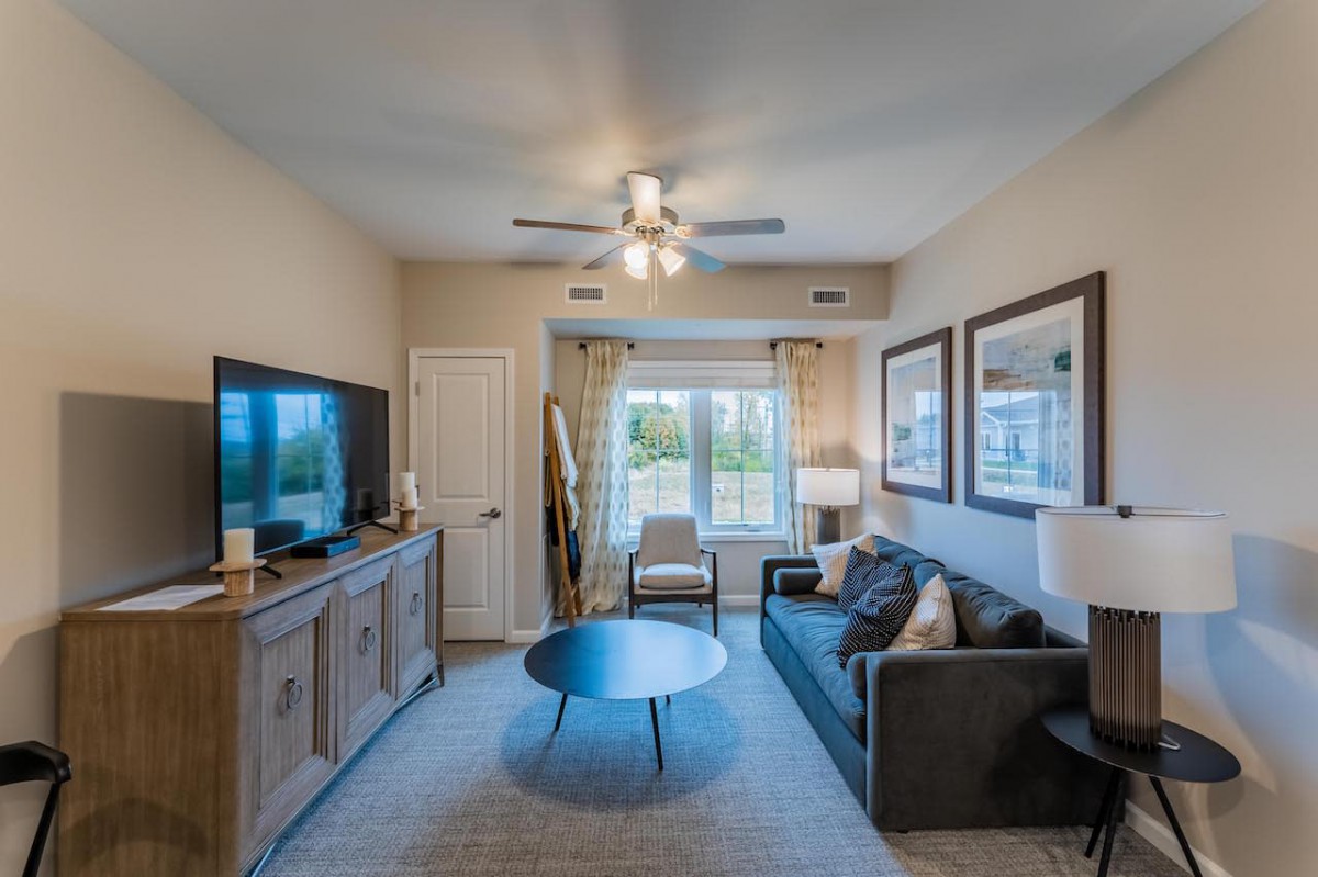 A cozy living room with a sofa, coffee table, and television, featuring a large window with curtains and a ceiling fan.