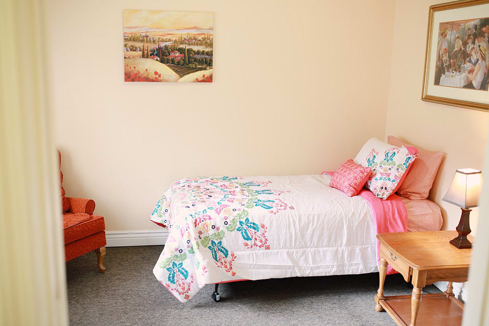 A cozy bedroom with a neatly made bed featuring a floral comforter and matching pillows, complemented by an orange chair and scenic wall art.