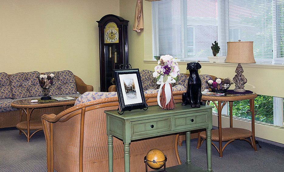 A cozy living room featuring a grandfather clock, wicker sofa, and a table with decorative items like a vase of flowers and a photo frame.
