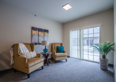 A cozy living room featuring two armchairs and a lamp, adorned with artwork, bathed in natural light from a sliding door.