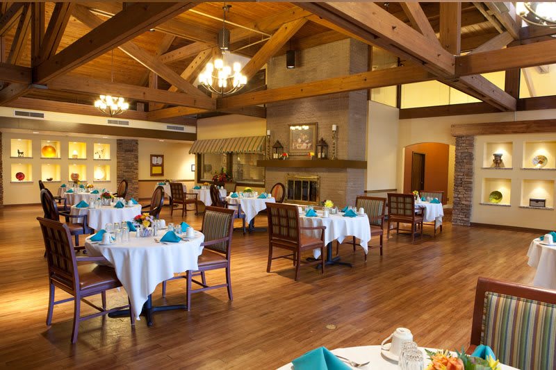 A cozy restaurant dining room with neatly arranged tables and chairs, wooden ceilings, and elegant chandeliers.