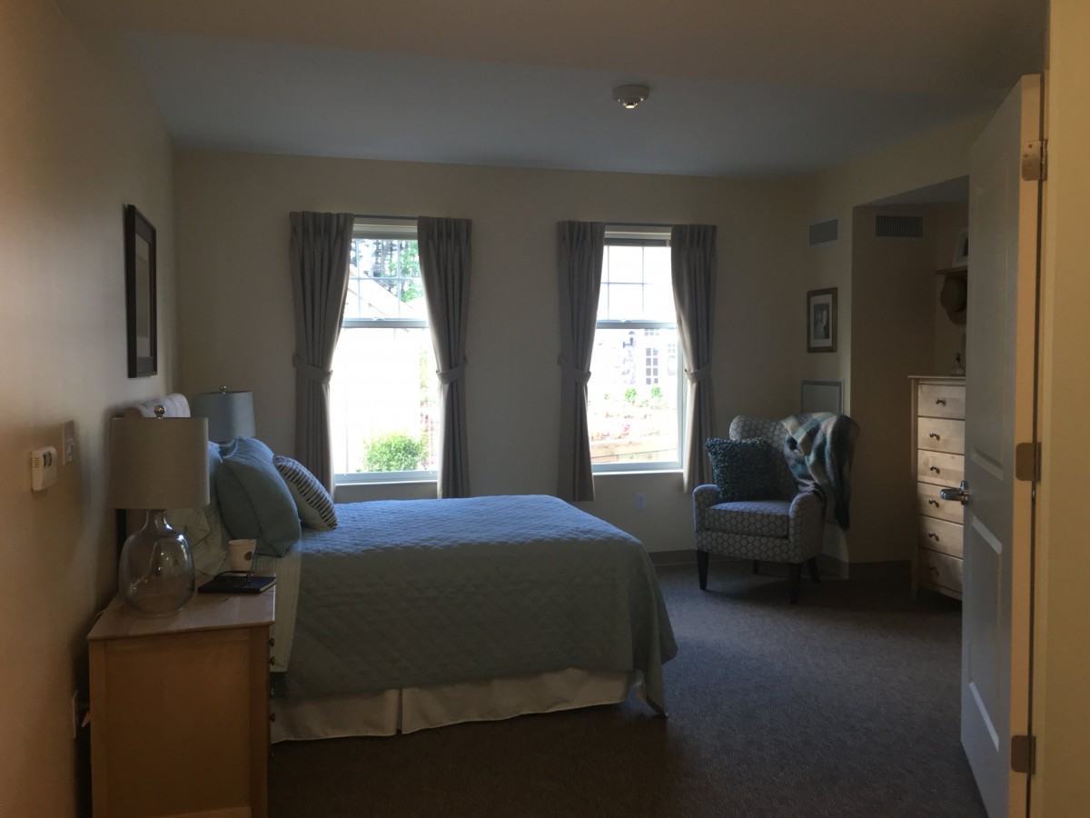 A cozy bedroom featuring a double bed, two windows with curtains, a comfortable chair, and a dresser.