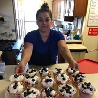 A woman presenting a tray of cupcakes topped with berries in a kitchen.