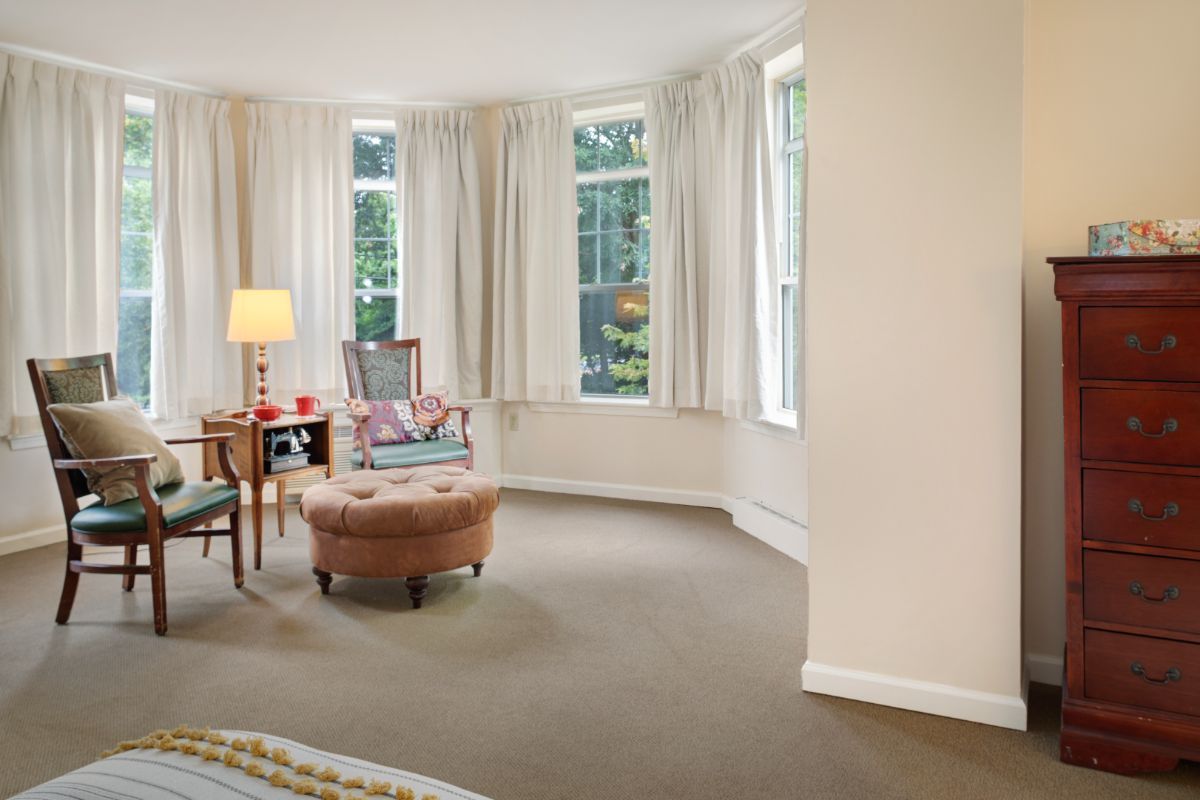 A cozy living room with an armchair, ottoman, and a lamp by large windows with curtains.