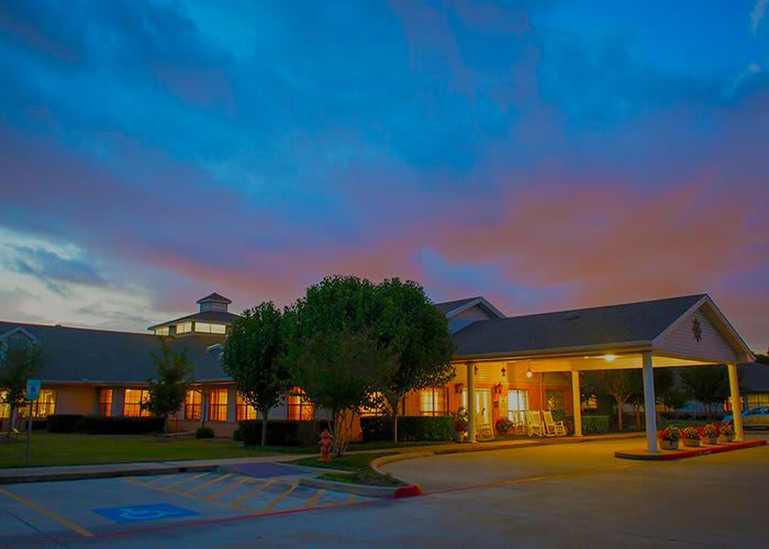 A warmly lit building with a parking area in the foreground, set against a colorful, cloudy evening sky.