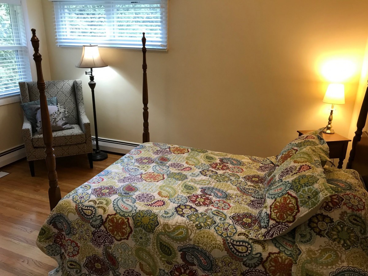A cozy bedroom with a colorful bedspread, featuring a lamp and a comfortable chair by the window.