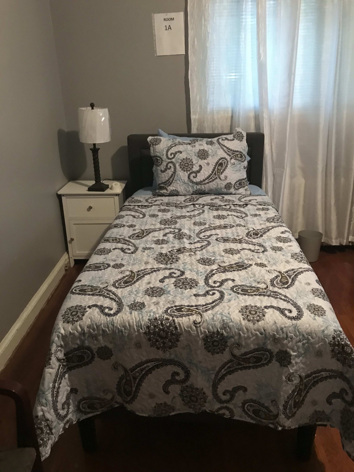 A cozy bedroom featuring a single bed with a paisley-patterned comforter, a nightstand with a lamp, and white curtains.