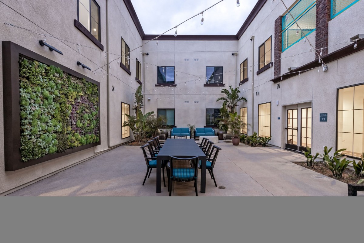 A modern courtyard with a long dining table, surrounded by plants and string lights.