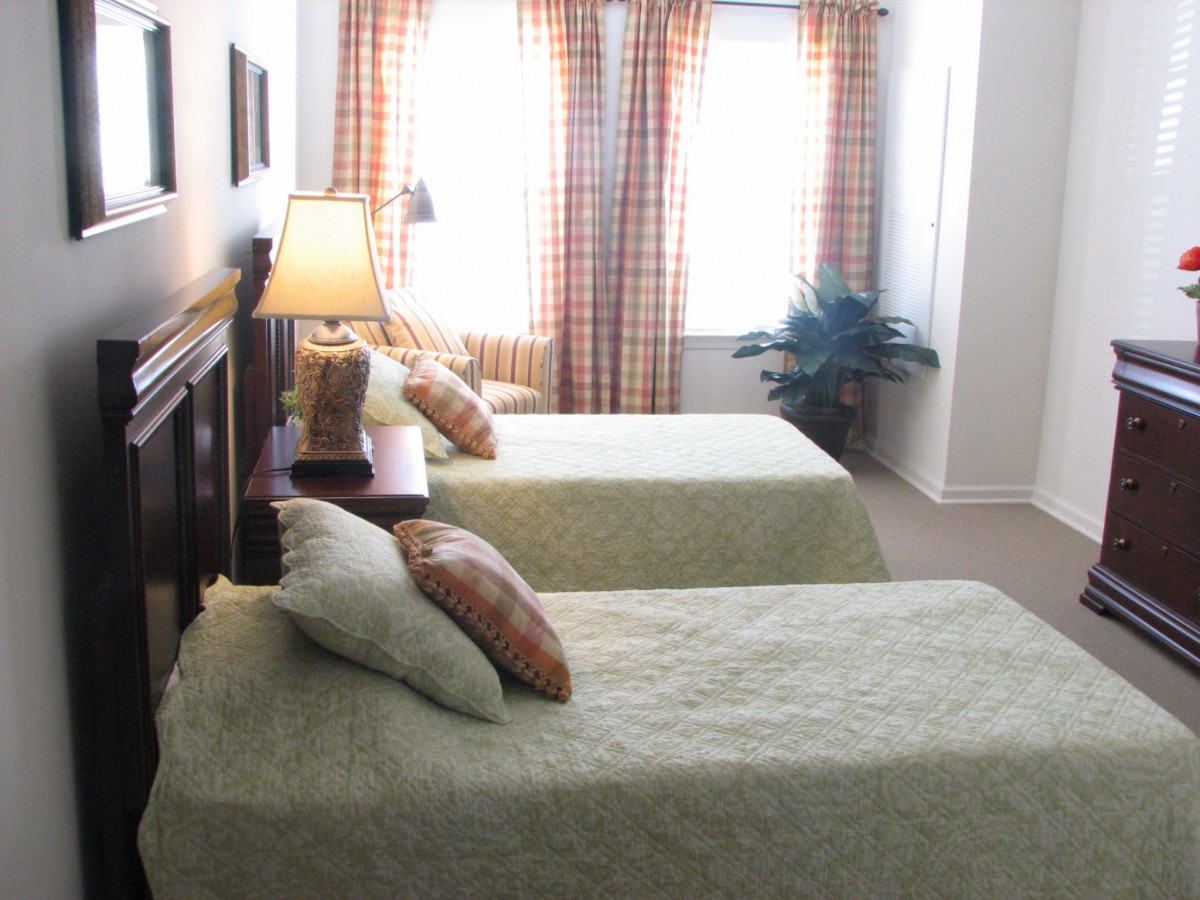 A cozy bedroom featuring twin beds with light green covers, a bedside lamp, and checkered curtains.