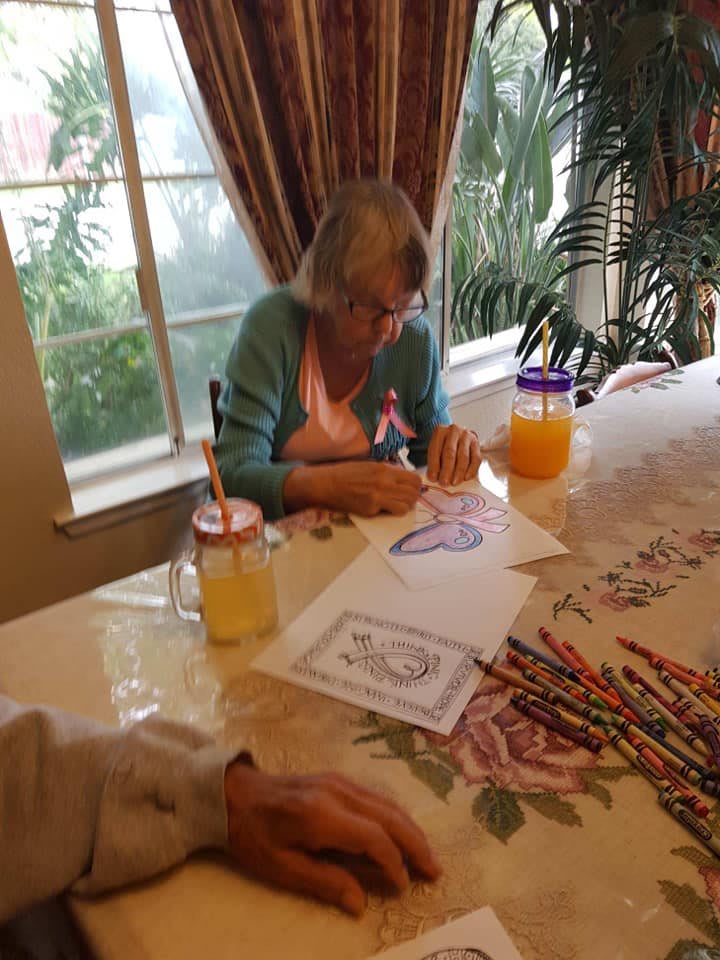 An elderly woman coloring at a table with art supplies and drinks.