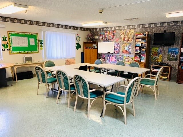 A community meeting room with arranged tables and chairs, featuring a notice board and bookshelves.