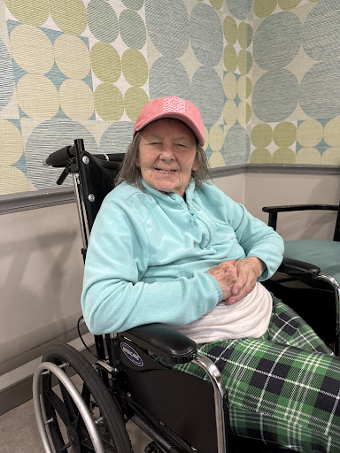A cheerful senior woman in a wheelchair, wearing a pink hat and light blue top, smiles warmly.