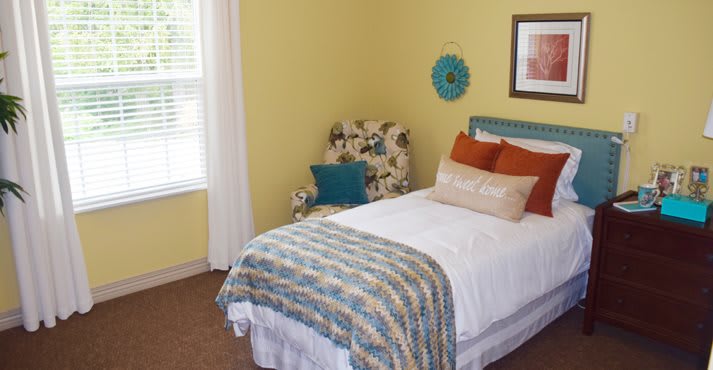 A cozy bedroom with a single bed, decorative pillows, and an armchair beside a window.