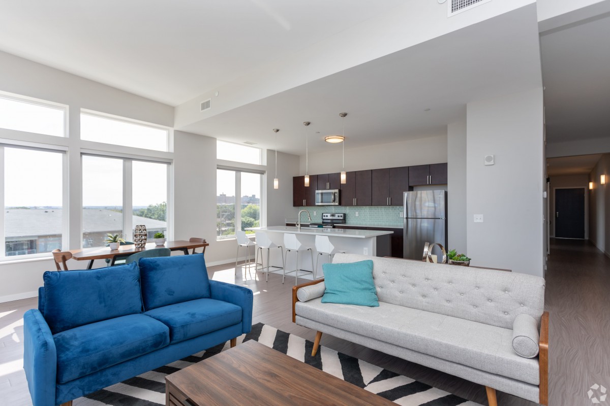 A modern living room with an open floor plan, featuring a blue sofa and natural light streaming in from large windows.