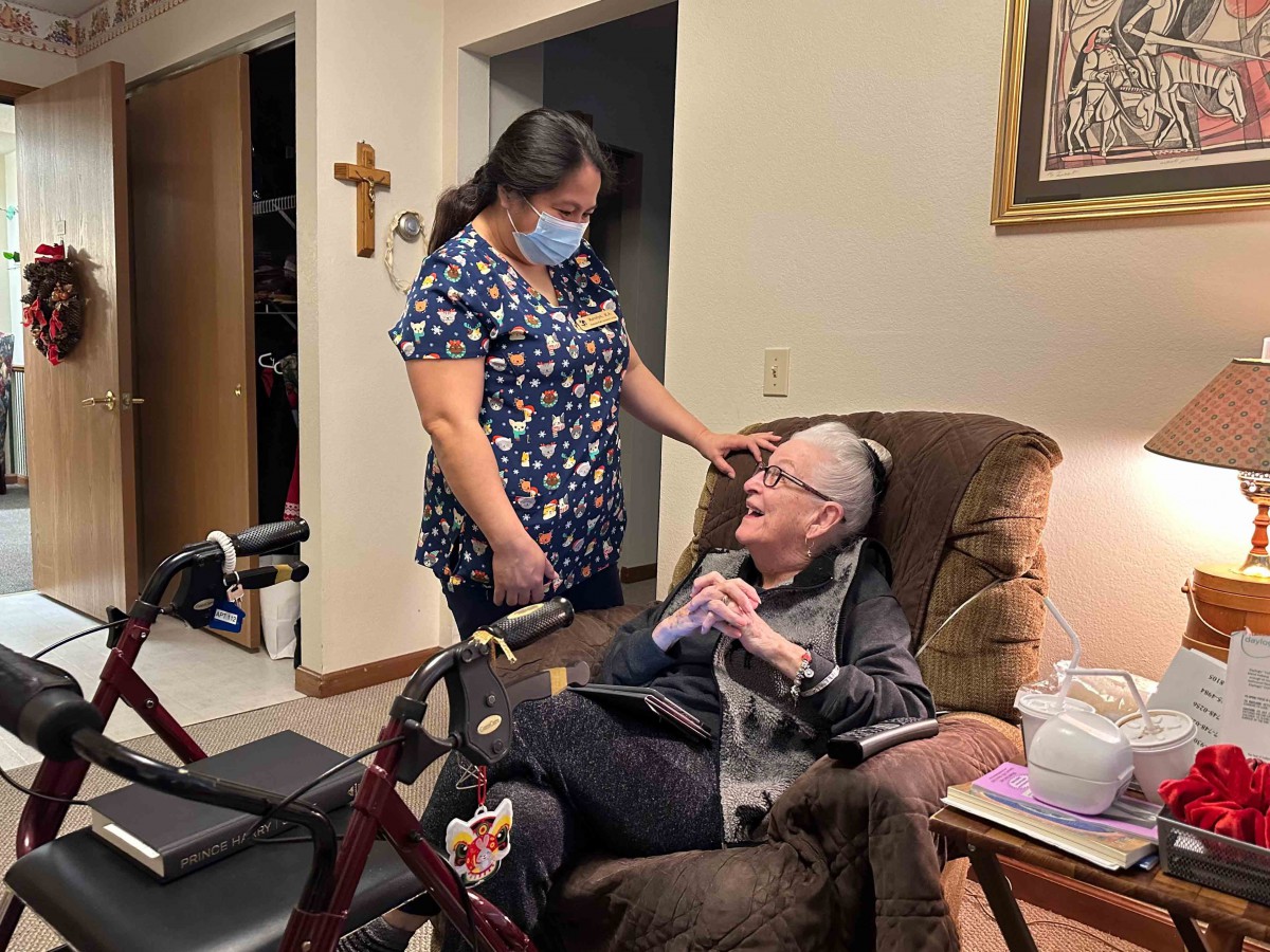 A caregiver wearing a mask interacts with an elderly person seated in a chair.