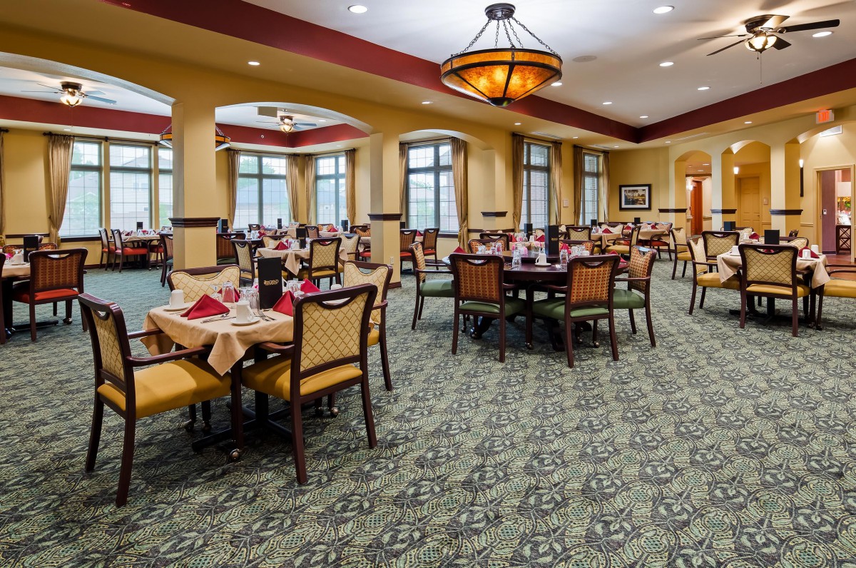 A spacious dining room with multiple tables and chairs, featuring elegant chandeliers and large windows.