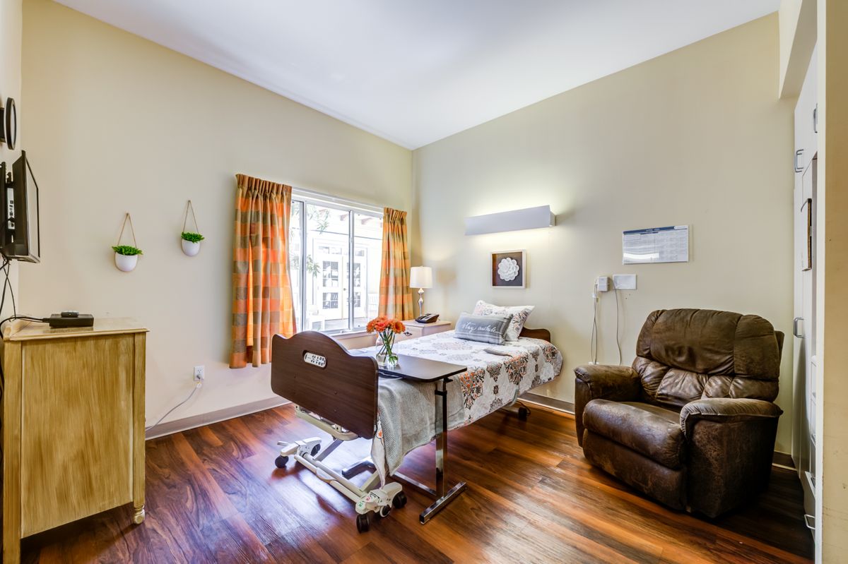 A well-lit hospital room with a patient bed and a recliner chair, featuring wooden floors and a large window with curtains.