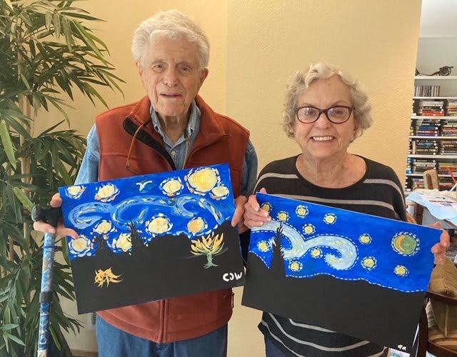 An elderly couple smiling while holding their Starry Night-inspired paintings indoors.