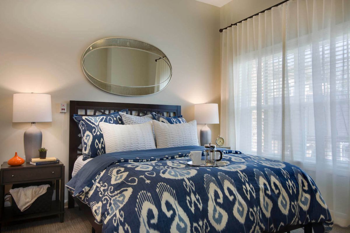 A cozy bedroom with a stylish blue and white patterned bedding set, complete with decorative pillows. An oval mirror hangs above the bed, complemented by a bedside lamp and sheer curtains letting in natural light.