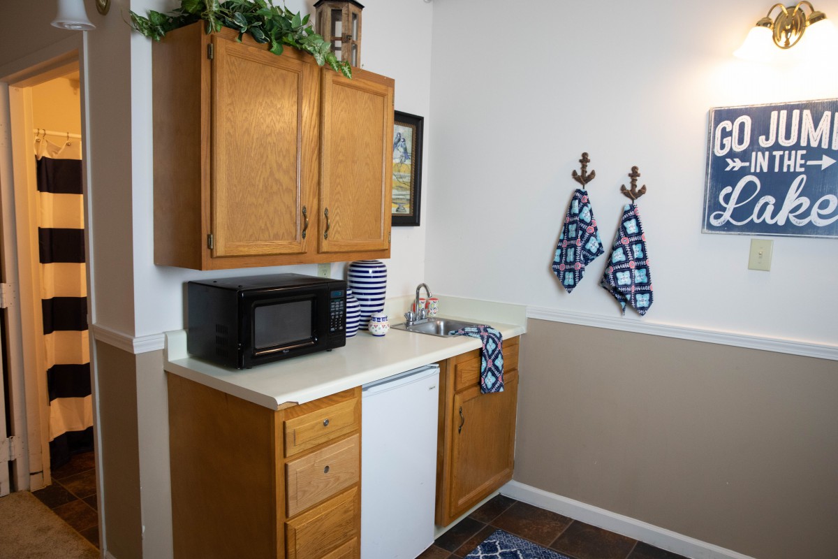 A small kitchen area with wooden cabinets, a countertop with a microwave, and decorative towels hanging on the wall.