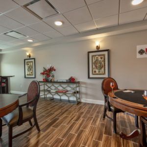A cozy room with wooden flooring, featuring tables and chairs, wall artwork, and warm lighting.