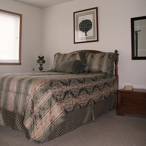 A cozy bedroom featuring a neatly made bed with patterned bedding, artwork on the wall, and a window allowing natural light to fill the room.