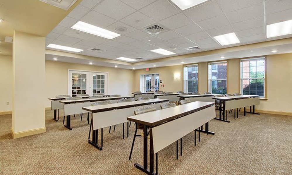 An empty classroom with rows of tables and chairs, bright lighting, and carpeted flooring.