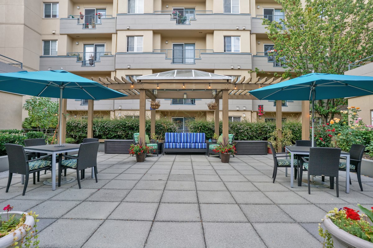 A modern apartment building with a spacious outdoor patio featuring pergolas, seating areas, and large umbrellas.