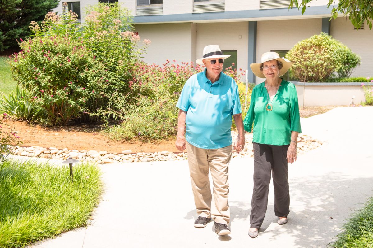An elderly couple walks together in a sunny garden, both wearing hats.