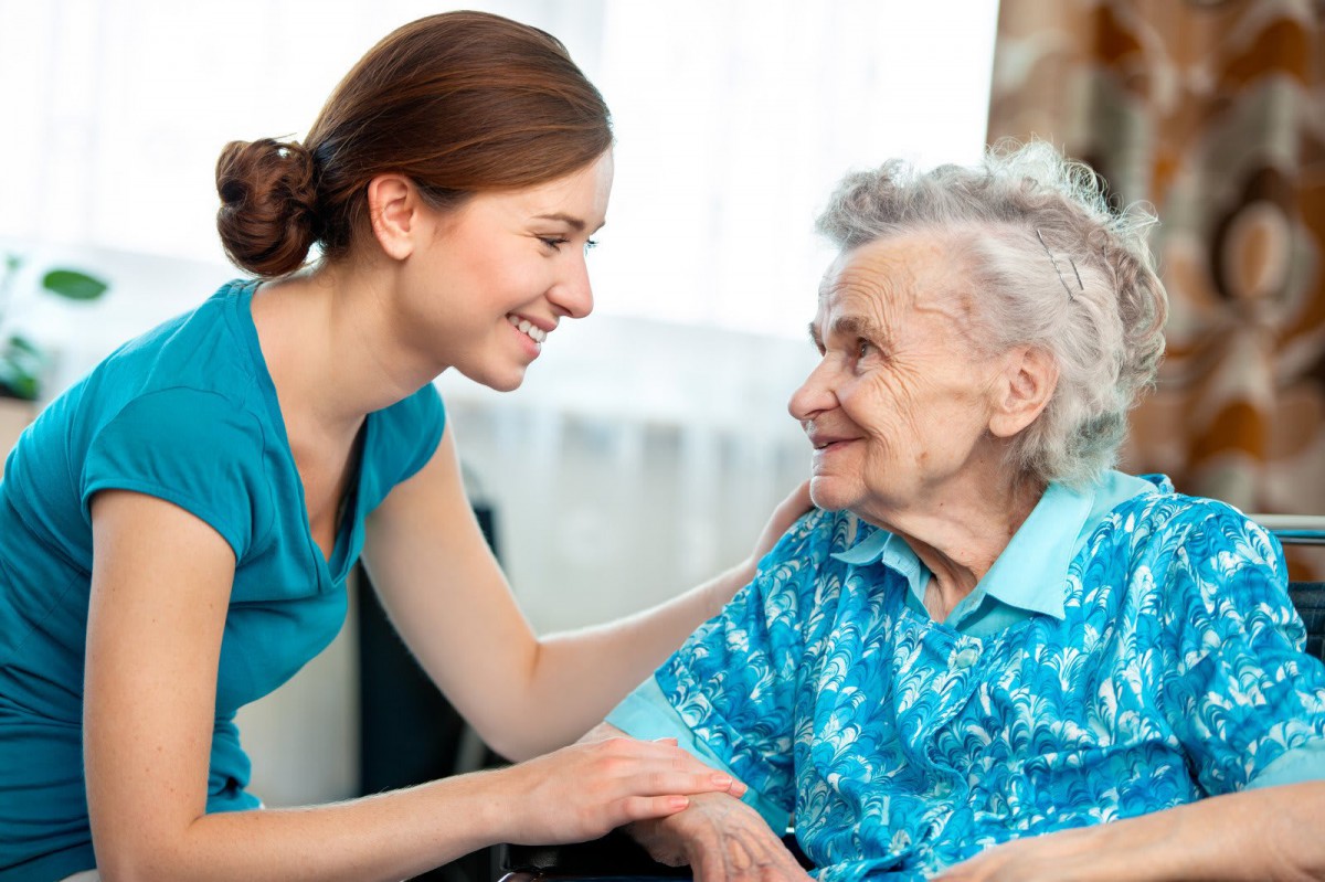 A caregiver smiles warmly at an elderly woman, showing support and companionship.